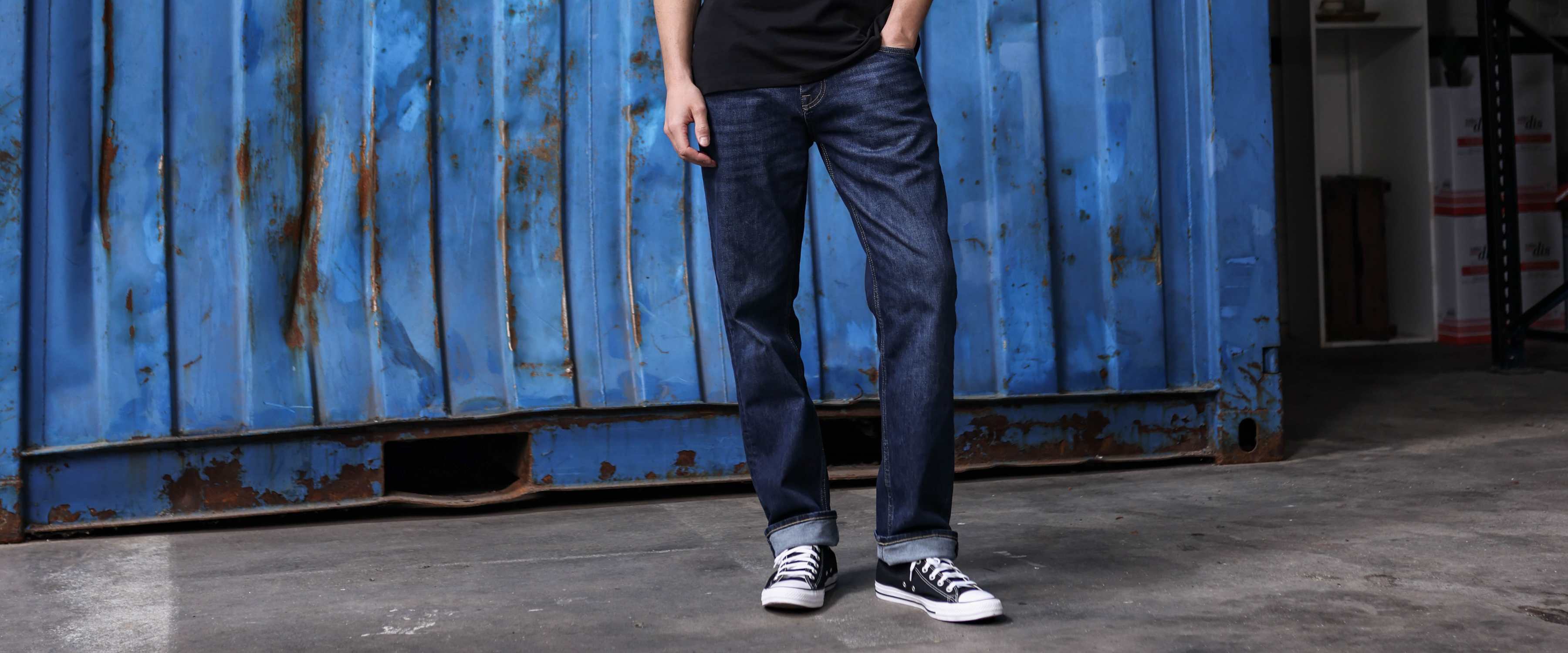 Een persoon in een donkerblauwe spijkerbroek, een zwart T-shirt en zwart-witte sneakers staat voor een verweerde blauwe scheepscontainer op een betonnen ondergrond.