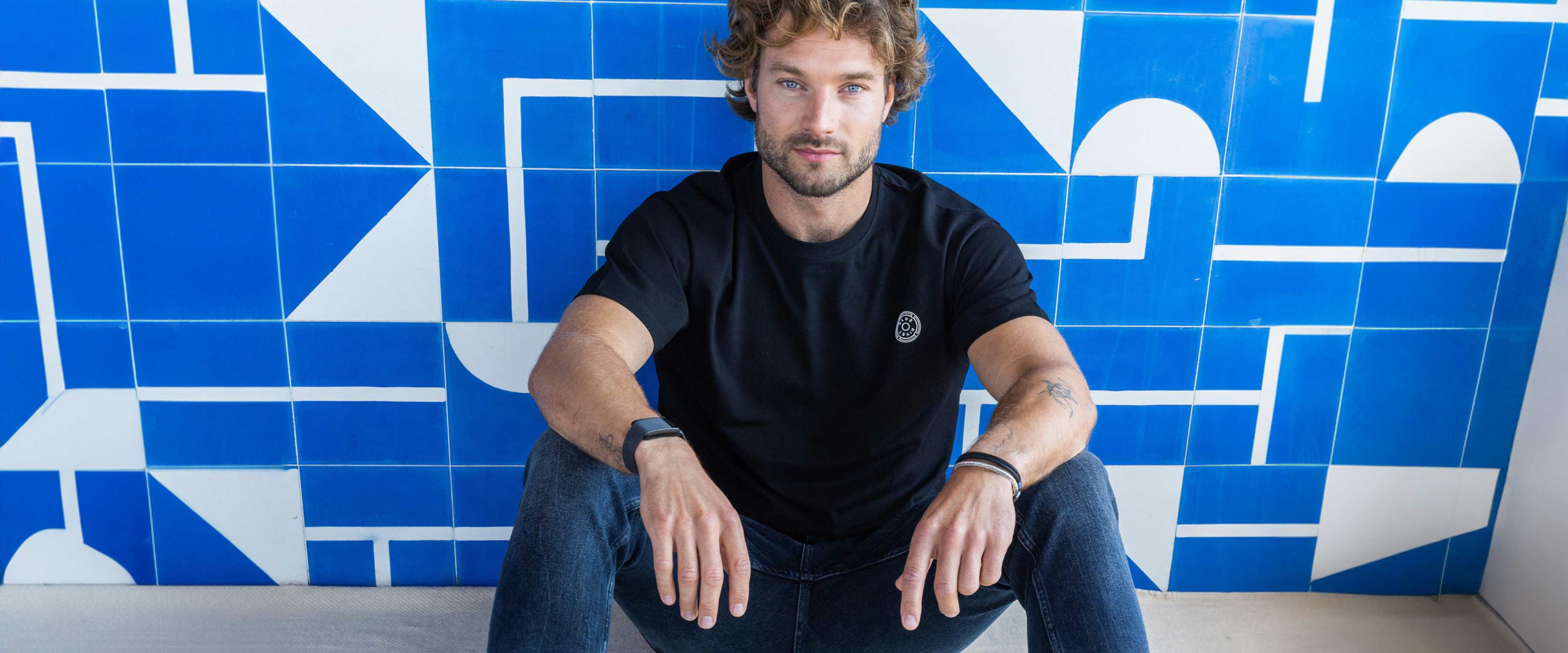 Een man met kort bruin haar en een baard, gekleed in een zwart T-shirt en een spijkerbroek, staat tegen een effen witte achtergrond, met zijn handen in zijn zakken en kijkt vooruit.