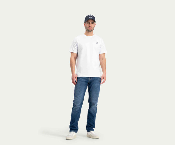 Een man staat met een wit T-shirt met een klein logo op de borst, de N01 Straight Indigo Used jeans van Blue Rivet Jeans, witte sneakers en een donkere baseballpet. De achtergrond is licht en effen.