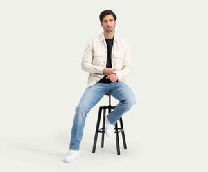 Een man met donker haar, gekleed in een lichtgekleurd jasje en The N01 Straight Stone Bleached denim jeans van Blue Rivet Jeans, zit op een zwarte kruk tegen een lichte achtergrond. Hij heeft een neutrale uitdrukking en kijkt vooruit, met zijn handen rustend op zijn knieën.