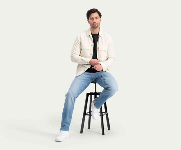 Een man met donker haar, gekleed in een lichtgekleurd jasje en The N01 Straight Stone Bleached denim jeans van Blue Rivet Jeans, zit op een zwarte kruk tegen een lichte achtergrond. Hij heeft een neutrale uitdrukking en kijkt vooruit, met zijn handen rustend op zijn knieën.