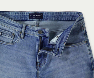Een close-up van de "The N01 Straight Stone Bleached" jeans benadrukt een gedeeltelijk open rits en knoop. Het straight-fit design heeft een label op de tailleband met de tekst Blue Rivet Jeans. De licht vervaagde, licht gedragen denim biedt zowel stijl als stretch voor comfort.