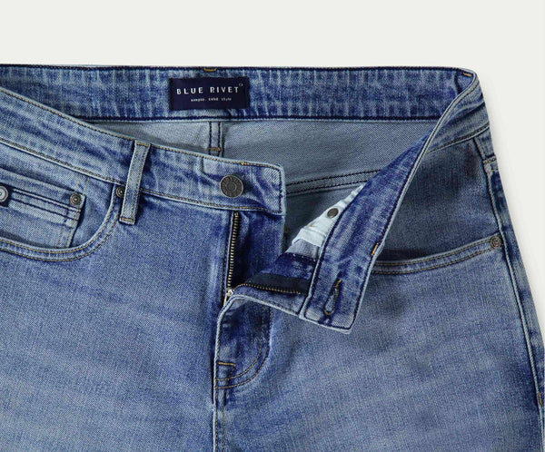 Een close-up van de "The N01 Straight Stone Bleached" jeans benadrukt een gedeeltelijk open rits en knoop. Het straight-fit design heeft een label op de tailleband met de tekst Blue Rivet Jeans. De licht vervaagde, licht gedragen denim biedt zowel stijl als stretch voor comfort.