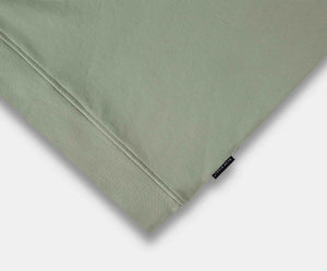De afbeelding toont een close-up van een gevouwen stof van The N01 Sweat Seagrass van Blue Rivet Jeans. Het is gemaakt van biologisch katoen in een lichtgroene kleur, met een klein zwart label met witte tekst aan de rechterrand. Het gladde maar licht gestructureerde materiaal belichaamt de kwaliteit van deze premium trui hier.
