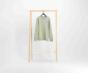 Een lichtgroene trui van Blue Rivet Jeans, bekend als The N01 Sweat Seagrass, gemaakt van biologisch katoen, hangt aan een zwarte hanger. Hij hangt op een houten rek tegen een effen witte achtergrond en heeft een normale pasvorm met een klein cirkelvormig ontwerp aan de linkerbovenkant.