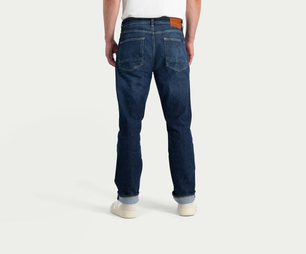 Een persoon staat met zijn rug naar de camera en draagt de N01 Straight Dark Indigo Used jeans van Blue Rivet Jeans, gestyled met opgerolde manchetten en een bruine leren patch op de tailleband. Ze worden gecombineerd met een strak wit shirt en witte sneakers tegen een effen lichtgrijze achtergrond.