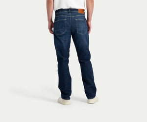 Een persoon is gekleed in Blue Rivet Jeans' "The N01 Straight Dark Indigo Used," gecombineerd met een wit T-shirt en witte sneakers. De jeans, van achteren gezien, benadrukken twee achterzakken en een bruine patch op de tailleband.