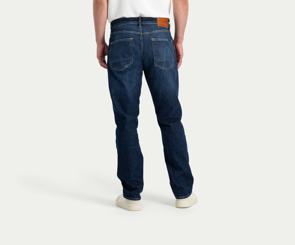 Een persoon is gekleed in Blue Rivet Jeans' "The N01 Straight Dark Indigo Used," gecombineerd met een wit T-shirt en witte sneakers. De jeans, van achteren gezien, benadrukken twee achterzakken en een bruine patch op de tailleband.