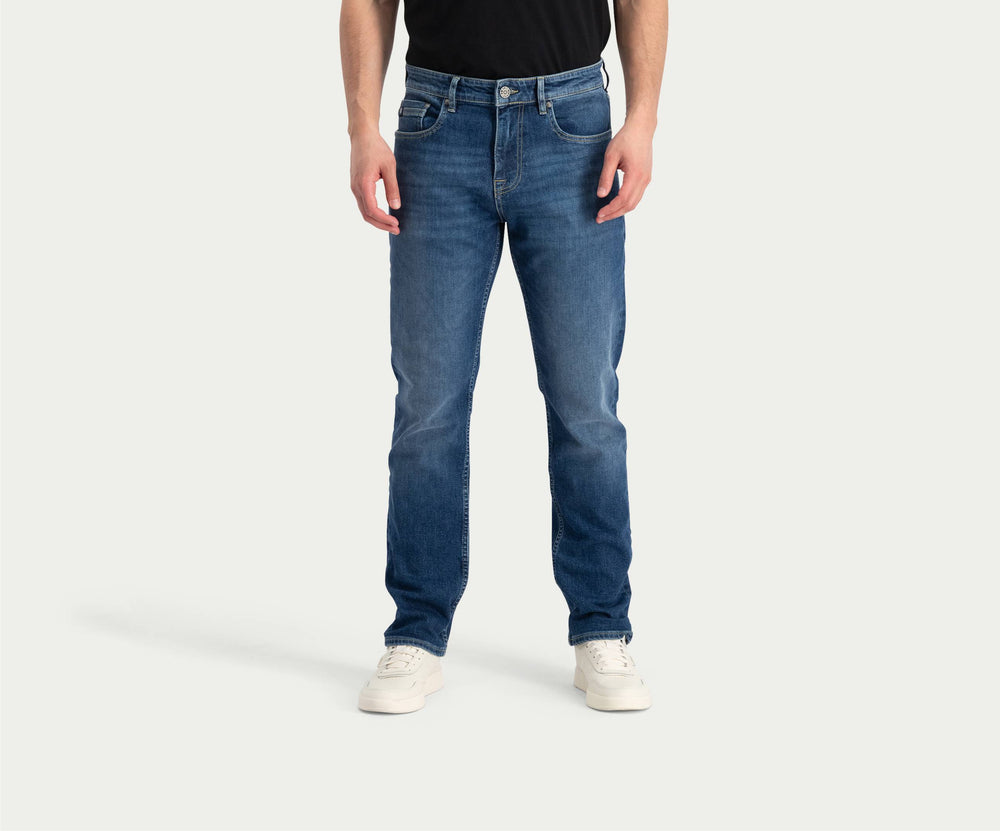 Een persoon die een zwart shirt draagt, de Blue Rivet Jeans' "The N01 Straight Indigo Used" met lichte vervaging, en witte sneakers staat tegen een effen achtergrond. Het hoofd van de persoon is niet zichtbaar op de afbeelding.