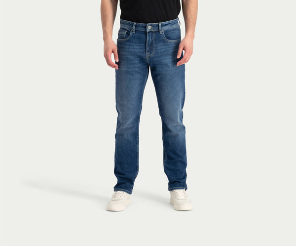Een persoon die een zwart shirt draagt, de Blue Rivet Jeans' "The N01 Straight Indigo Used" met lichte vervaging, en witte sneakers staat tegen een effen achtergrond. Het hoofd van de persoon is niet zichtbaar op de afbeelding.
