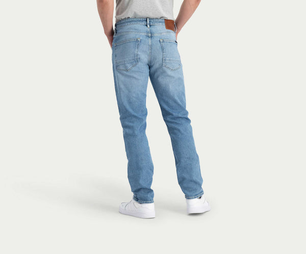 Een persoon is van achteren te zien met de N01 Straight Stone Bleached jeans van Blue Rivet Jeans en witte sneakers. De lichtblauwe denim jeans heeft zichtbare achterzakken en een bruine leren patch op de tailleband, allemaal tegen een effen witte achtergrond.