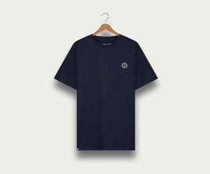 Een marineblauwe The N01 Tee van Blue Rivet Jeans hangt aan een houten hanger tegen een lichte achtergrond. Dit regular fit shirt heeft een klein rond embleem op de linkerborst en een contrasterend halsdetail.