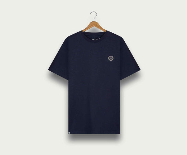 Een marineblauwe The N01 Tee van Blue Rivet Jeans hangt aan een houten hanger tegen een lichte achtergrond. Dit regular fit shirt heeft een klein rond embleem op de linkerborst en een contrasterend halsdetail.