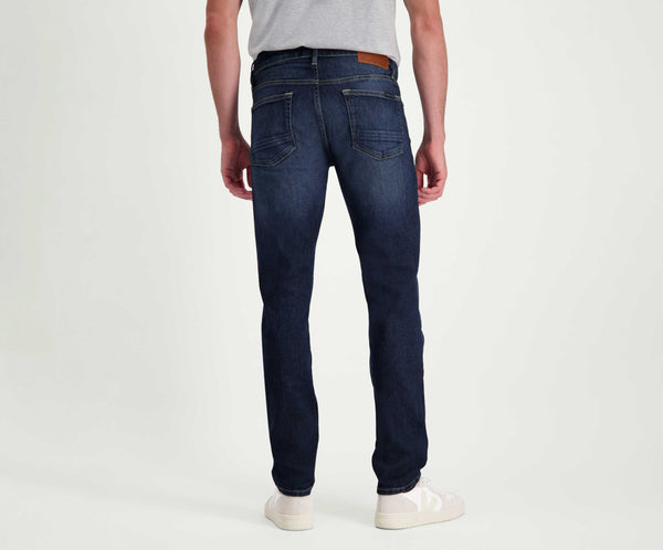 Een persoon die een grijs t-shirt en The N01 Slim Dark Indigo Used jeans van Blue Rivet Jeans draagt, staat met zijn rug naar de camera en laat de premium pasvorm en het design van zijn jeans zien. De afbeelding benadrukt de vorm van deze spijkerbroek, aangevuld met witte sneakers.