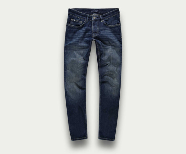 De N01 Slim Dark Indigo Used jeans van Blue Rivet Jeans zijn gemaakt van stretch denim en hebben een knoop- en ritssluiting. Deze premium jeans hebben subtiele vervagingen op de dijen en knieën, vijf zakken en contrasterende stiksels, allemaal tegen een lichtgrijze achtergrond.