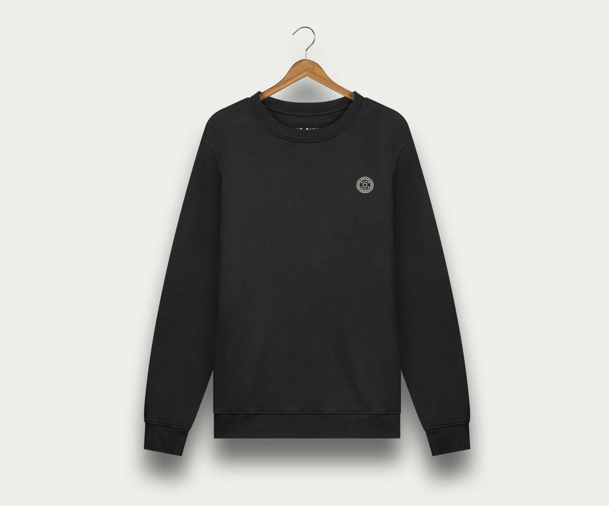 De N01 Sweat Anthracite van Blue Rivet Jeans is een zwarte crewneck sweater van biologisch katoen, afgebeeld op een houten hanger tegen een lichte achtergrond. Deze premium sweater voor heren heeft een klein rond logo op de linkerborst.