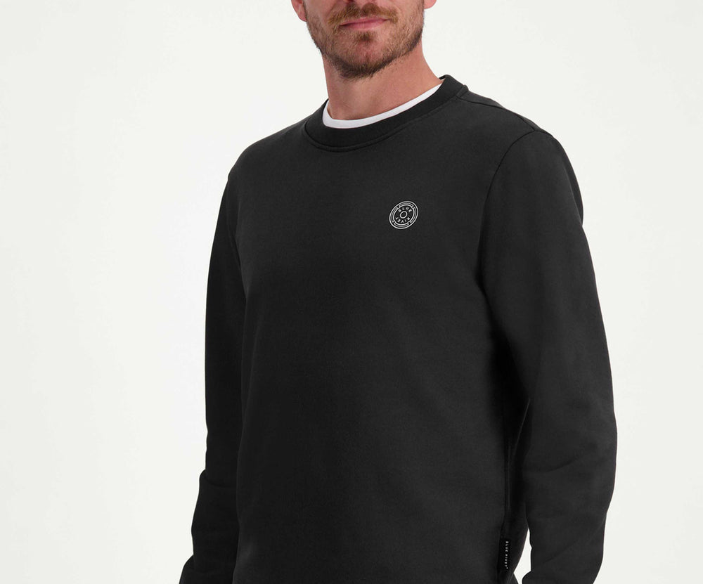 Een man met kort bruin haar en een baard draagt The N01 Sweat Anthracite van het merk Blue Rivet Jeans, gecombineerd met een regular fit blauwe jeans. Hij staat tegen een effen witte achtergrond en kijkt lichtjes naar links. De sweater heeft een klein rond logo op de borst.
