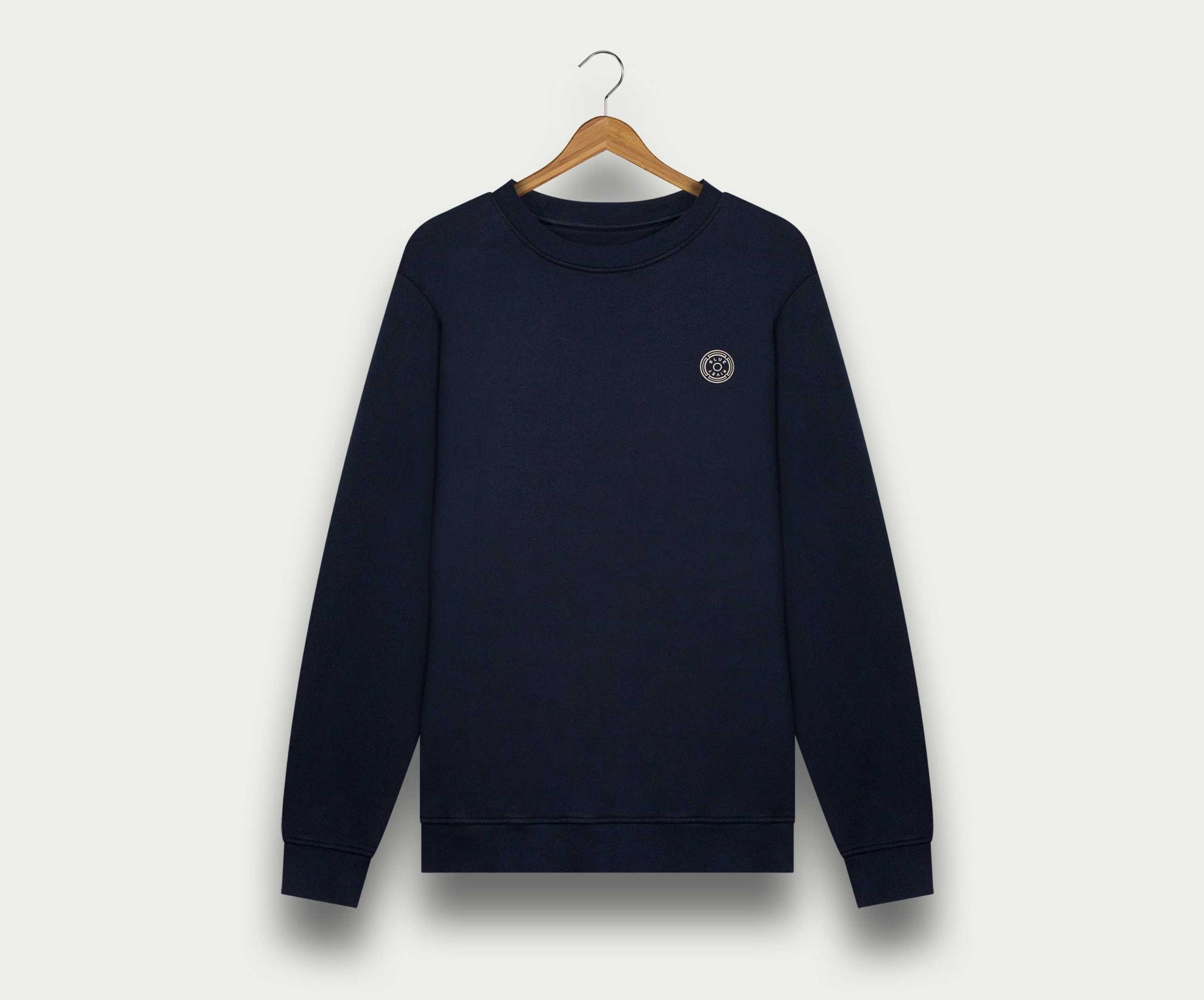 Een marineblauwe herentrui met lange mouwen van Blue Rivet Jeans, gemaakt van biologisch katoen, heeft een klein rond logo op de linkerborst. Hij hangt elegant aan een houten hanger tegen een effen, lichtgekleurde achtergrond, wat de ingetogen stijl benadrukt.
