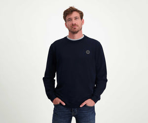 Een man met kort haar en een baard draagt The N01 Sweat Navy, een donkere, regular fit, long-sleeve trui van Blue Rivet Jeans. Hij is gemaakt van biologisch katoen en heeft een klein rond logo op de borst. Hij heeft hem gecombineerd met jeans en staat nonchalant met zijn handen in zijn zakken tegen een effen witte achtergrond.