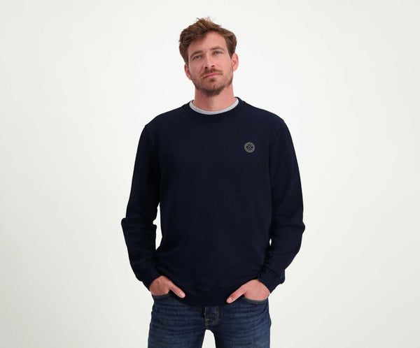 Een man met kort haar en een baard draagt The N01 Sweat Navy, een donkere, regular fit, long-sleeve trui van Blue Rivet Jeans. Hij is gemaakt van biologisch katoen en heeft een klein rond logo op de borst. Hij heeft hem gecombineerd met jeans en staat nonchalant met zijn handen in zijn zakken tegen een effen witte achtergrond.