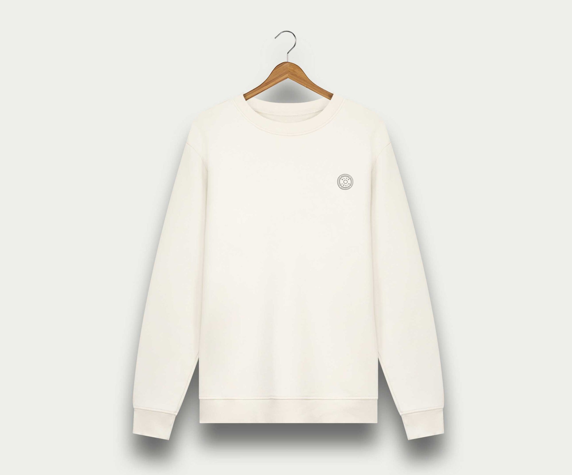 Aan een houten hanger tegen een witte achtergrond hangt The N01 Sweat Off White van Blue Rivet Jeans, een effen witte herentrui met een klein rond logo op de linkerborst en gemaakt van biologisch katoen.