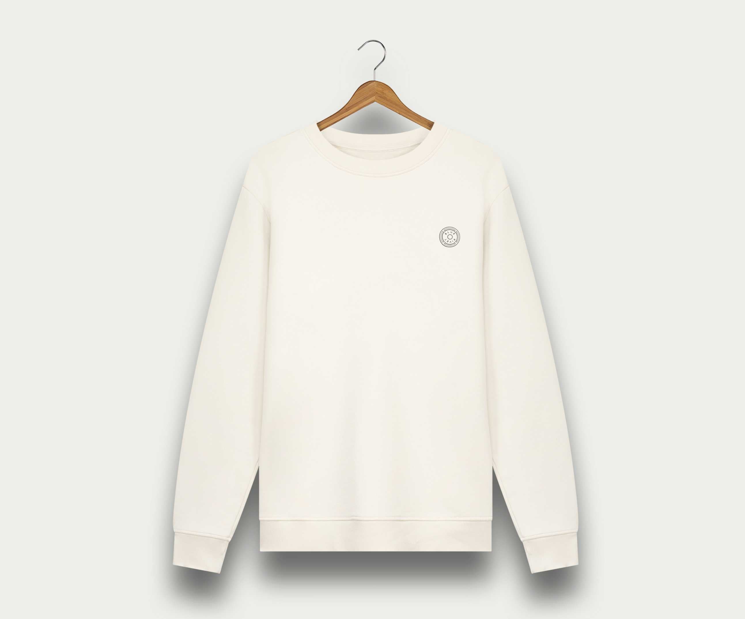 Aan een houten hanger tegen een witte achtergrond hangt The N01 Sweat Off White van Blue Rivet Jeans, een effen witte herentrui met een klein rond logo op de linkerborst en gemaakt van biologisch katoen.