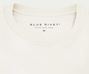 Close-up van een off-white sweatshirt met het bovenste gedeelte. Het label vermeldt Blue Rivet Jeans en een eenvoudige. effen. stijl. met een maat M. De N01 Sweat Off White is gemaakt van biologisch katoen en heeft een zachte stof en een minimalistisch design, perfect voor degenen die ingetogen elegantie waarderen.