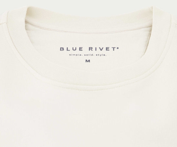 Close-up van een off-white sweatshirt met het bovenste gedeelte. Het label vermeldt Blue Rivet Jeans en een eenvoudige. effen. stijl. met een maat M. De N01 Sweat Off White is gemaakt van biologisch katoen en heeft een zachte stof en een minimalistisch design, perfect voor degenen die ingetogen elegantie waarderen.