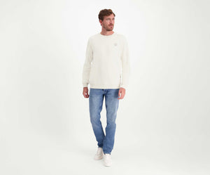 Een persoon met kort bruin haar draagt de N01 Sweat Off White trui van Blue Rivet Jeans, gecombineerd met een blauwe jeans en witte sneakers. Hij staat tegen een effen witte achtergrond en kijkt opzij.