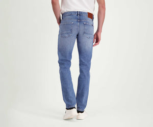 Een achteraanzicht van een persoon die Blue Rivet Jeans' The N01 Slim Stone Bleached draagt, gecombineerd met een wit t-shirt en witte sneakers, staand tegen een effen witte achtergrond. De jeans hebben een leren patch op de tailleband.
