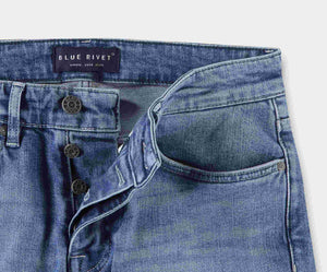 Een close-up van een paar The N01 Slim Stone Bleached jeans met een gedeeltelijk open knoopsluiting. Het Blue Rivet Jeans-label is prominent aanwezig, terwijl de denim zichtbare stiksels en zakdetails heeft, versterkt door de stretchstof. Deze spijkerbroek tegen een witte achtergrond straalt zowel stijl als comfort uit.