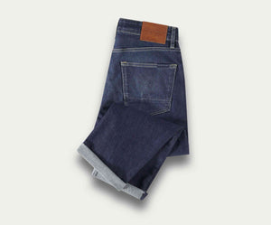 Een netjes opgevouwen paar The N01 Straight Indigo Used jeans van Blue Rivet Jeans, met een bruine leren patch op de tailleband en een zichtbare achterzak, geplaatst op een lichte achtergrond.