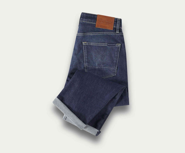Een netjes opgevouwen paar The N01 Straight Indigo Used jeans van Blue Rivet Jeans, met een bruine leren patch op de tailleband en een zichtbare achterzak, geplaatst op een lichte achtergrond.