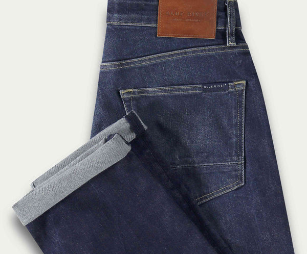 De N01 Straight Indigo Used van Blue Rivet Jeans wordt netjes gevouwen weergegeven met een omgeslagen pijp, met een leren patch op de tailleband met tekst en een achterzak. De licht versleten textuur van de stretchdenim combineert stijl met comfort.