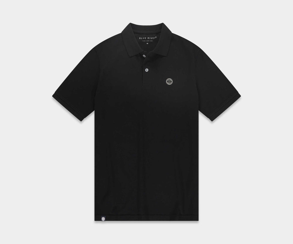 Een man zit op een zwarte kruk tegen een effen beige achtergrond, en straalt zomerse chic uit. Hij draagt The N01 Polo Anthracite, een klassieke polo van biologisch katoen, gecombineerd met de blauwe denim van Blue Rivet Jeans en stijlvolle zwarte sneakers. Met zijn armen over elkaar kijkt hij recht in de camera, wat een moeiteloze stijl uitstraalt.