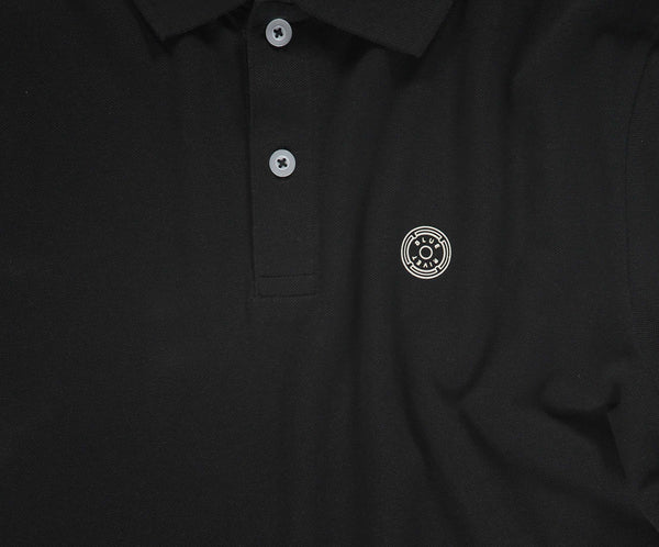 De N01 Polo Anthracite van Blue Rivet Jeans is een zwarte piqué polo, afgewerkt met grijze knopen en versierd met een cirkelvormig embleem met een abstract ontwerp op de linkerborst. Het is gemaakt van biologisch katoen voor een vleugje zomerse chic.