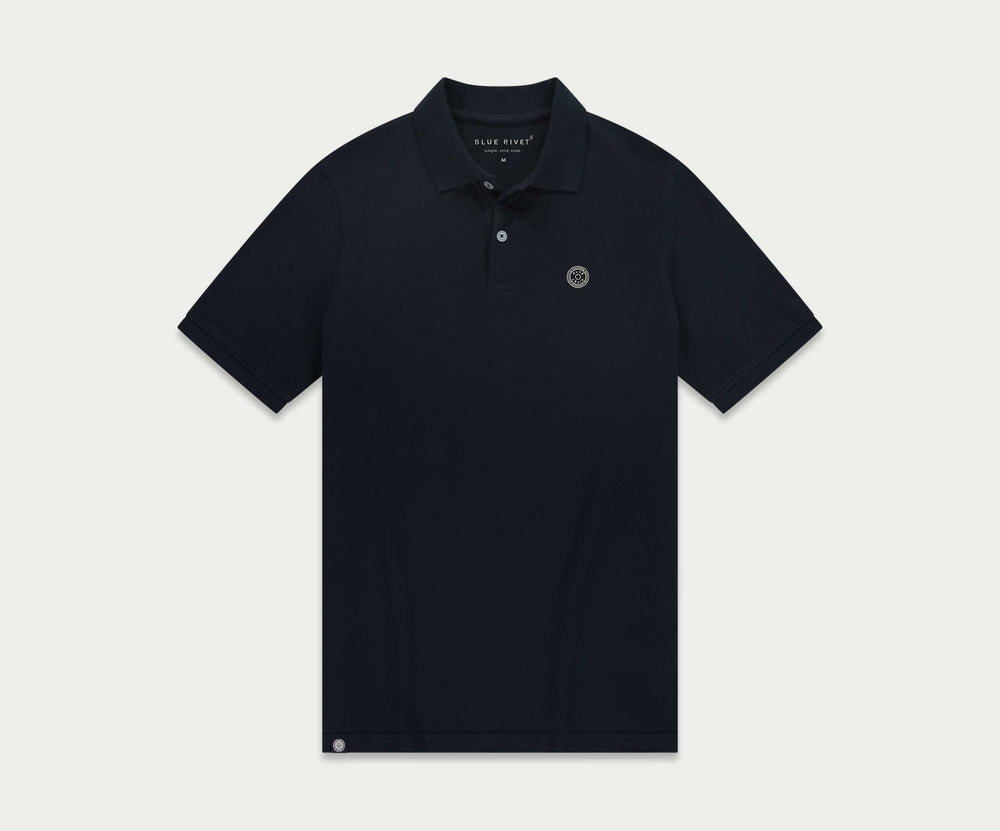 Tegen een effen beige achtergrond is een man te zien in The N01 Polo Navy en lichtblauwe Blue Rivet Jeans, aangevuld met witte sneakers. Hij heeft kort donker haar en een neutrale uitdrukking.