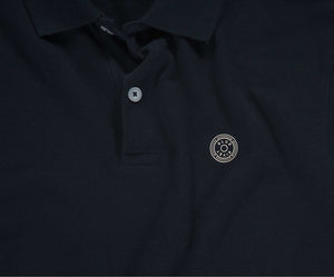 De marineblauwe N01 Polo van Blue Rivet Jeans heeft een kraag met knopen en een gestileerd zilveren cirkelvormig embleem op de linkerborst. De polo heeft korte mouwen.