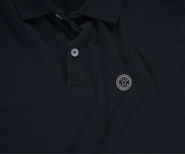 De marineblauwe N01 Polo van Blue Rivet Jeans heeft een kraag met knopen en een gestileerd zilveren cirkelvormig embleem op de linkerborst. De polo heeft korte mouwen.