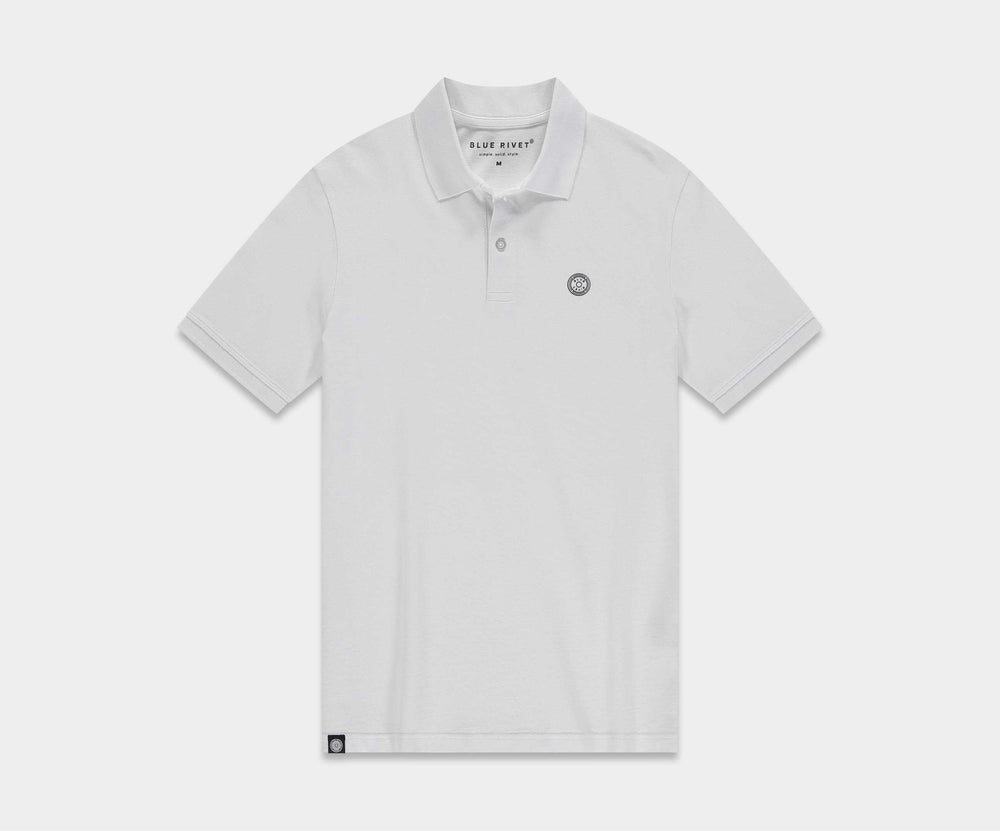 Een man die de N01 Polo White en grijze Blue Rivet Jeans draagt, samen met witte sneakers, zit op een kruk tegen een lichtbeige achtergrond. Hij lijkt ontspannen met één voet op de vloer en zijn handen rustend op zijn schoot.
