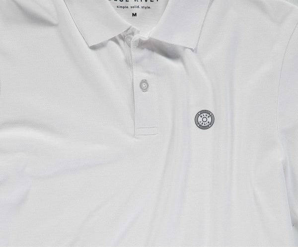 Een wit poloshirt van Blue Rivet Jeans, bekend als The N01 Polo White, met korte mouwen en een klein cirkelvormig embleem met patroon op de linkerborst. Dit shirt heeft een kraag en twee knopen. Binnenin staat op het label BLUE RIVER simple. solid. style. M.