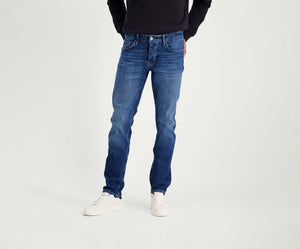 Een persoon die Blue Rivet Jeans' The N01 Slim Indigo Used draagt, staat tegen een effen achtergrond. Hun handen zitten in de zakken van de jeans en het bovenlichaam is gedeeltelijk zichtbaar, gekleed in een strak zwart topje, aangevuld met witte sneakers.