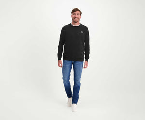 Een man staat in een ontspannen houding, draagt een zwarte trui, The N01 Slim Indigo Used jeans van Blue Rivet Jeans en witte sneakers. De achtergrond is effen wit. Hij heeft een lichte glimlach en zijn handen zijn nonchalant langs zijn lichaam.