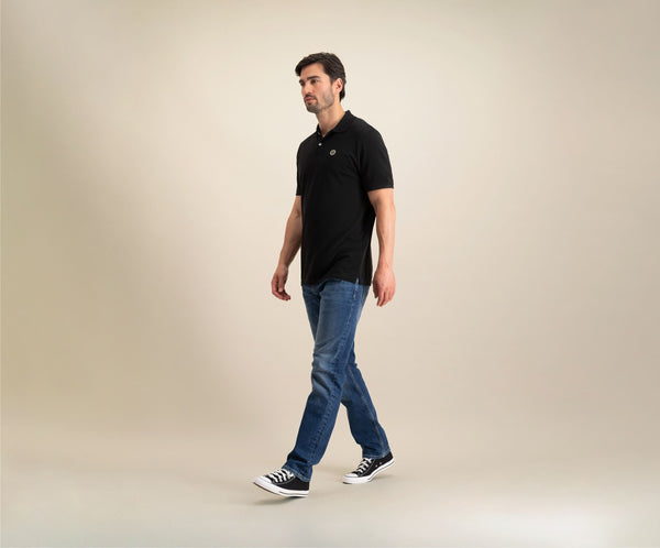 Een man die The N01 Polo Anthracite en Blue Rivet Jeans draagt, loopt zelfverzekerd over een effen, zacht verlichte achtergrond. Met zwarte en witte sneakers straalt hij zomerse chic uit terwijl hij met een ontspannen houding naar links kijkt.