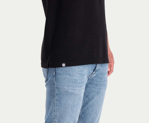 Een persoon die The N01 Polo Anthracite and Blue Rivet Jeans draagt, in een lichtblauwe wassing, staat tegen een effen achtergrond. Alleen het onderste deel van het poloshirt en de jeans met een klein label zichtbaar op het shirt worden getoond, wat een vleugje zomerse chic toevoegt aan hun ingetogen look.