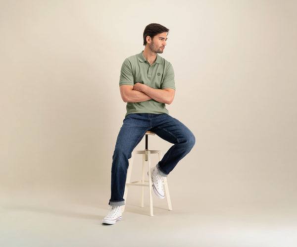 Een man zit op een hoge witte kruk tegen een neutrale achtergrond, draagt The N01 Polo Seagrass met korte mouwen. Zijn armen zijn gekruist terwijl hij opzij kijkt, gecombineerd met Blue Rivet Jeans en witte sneakers.