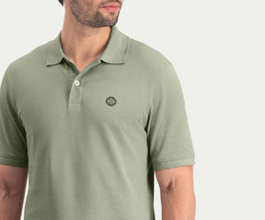 Een man draagt The N01 Polo Seagrass van Blue Rivet Jeans, met korte mouwen en een klein rond logo op de linkerborst. De afbeelding is bijgesneden op de schouders en taille, met de nadruk op de details van het shirt.