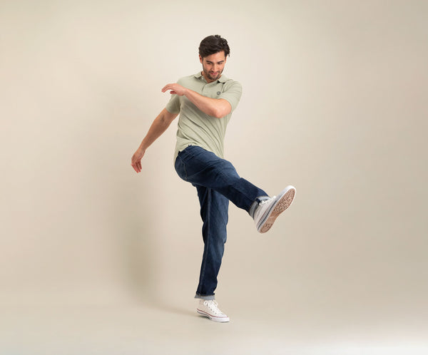 Een man met kort donker haar, gekleed in de N01 Polo in zeegras, een blauwe Rivet-spijkerbroek en witte sneakers, is halverwege zijn pas en schopt zijn been naar voren tegen een effen beige achtergrond.