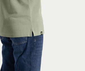 Een persoon is gedeeltelijk zichtbaar, draagt The N01 Polo Seagrass met korte mouwen en Blue Rivet Jeans. Het shirt heeft een kleine zijsplit met een merklabel, terwijl de jeans een klassieke blauwe denimtextuur hebben, allemaal tegen een effen lichte achtergrond.