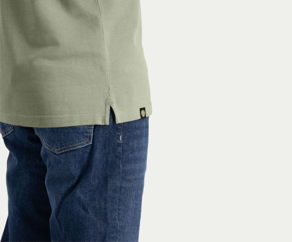 Een persoon is gedeeltelijk zichtbaar, draagt The N01 Polo Seagrass met korte mouwen en Blue Rivet Jeans. Het shirt heeft een kleine zijsplit met een merklabel, terwijl de jeans een klassieke blauwe denimtextuur hebben, allemaal tegen een effen lichte achtergrond.